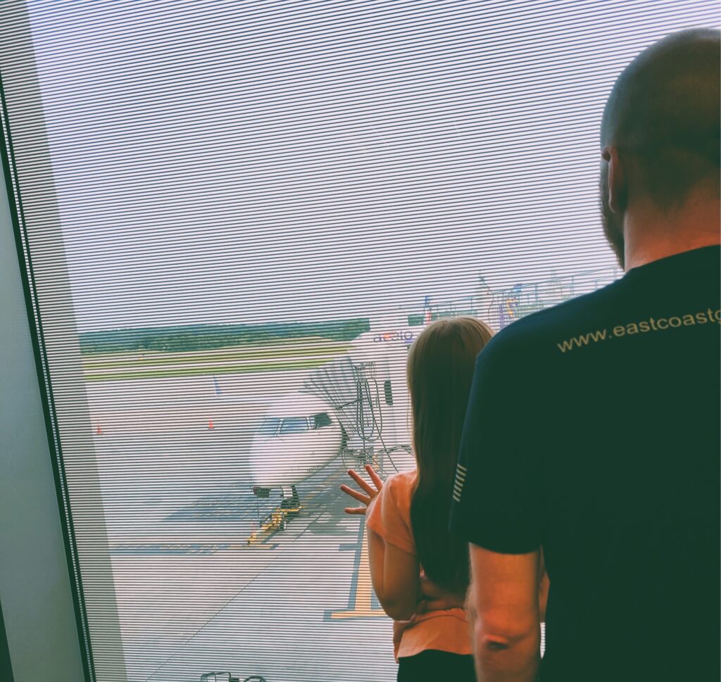 Photo of a girl and dad looking out an airport window at a plane.